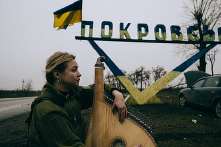 A singer with a stringed instrument near the Provosk city sign (in Cyrillic lettering), to which is attached a tattered Ukraine flag