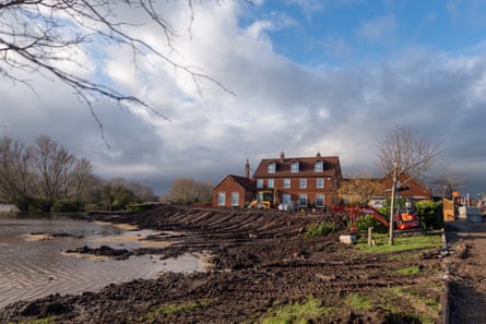 A red and a yellow digger move earth to form a muddy brown bank around a large detached brick house close to the river.