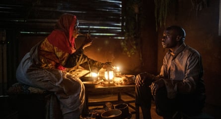 A woman smokes a pipe at a low table in a dark hut lit by a lamp as a man sits opposite her