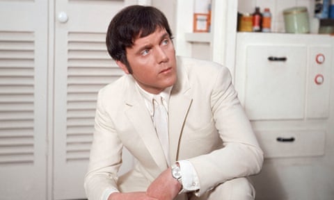 A young Kenneth Cope in a white suit, kneeling on the ground in a white-furnished room