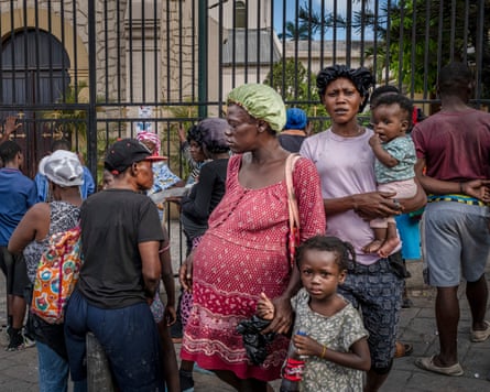 A pregnant woman holds the hand of a child in a crowd of people in front of the gates of what appears to be a church