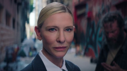Headshot of Cate Blanchett in a shirt and dark suit, with a dishevelled man blurred in the background on a city street