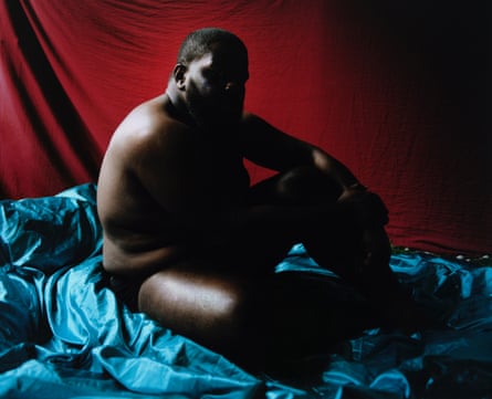 A man siting on a blue drape in underwear, his face in shadow, clutching one knee
