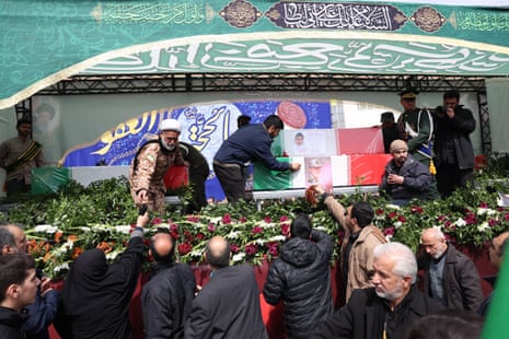 लोगों का एक समूह एक मंच के सामने इकट्ठा होता है, जहाँ वे गुलाब और अन्य फूल बिछा रहे हैं।