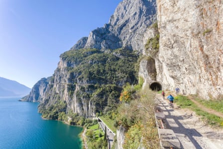 Cyclists approach a tunnel in rock cliffs by a lake on sunny day