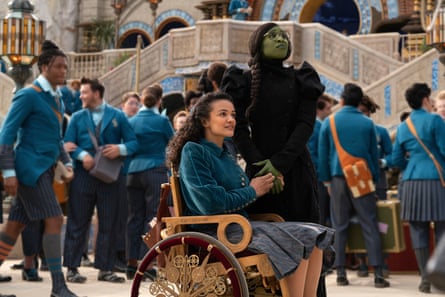 Marissa Bode, wearing a blue and grey school uniform, seated in a wheelchair, and Cynthia Erivo, standing, in green face makeup and long black garb, in a school setting as sisters Nessarose and Elphaba.