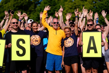 Elizabeth Duggan, chief executive officer of Parkrun, Parkrun founder, Paul Sinton-Hewitt and Dame Kelly Holmes at the Bushy Park Parkrun in London, where the very first Parkrun took place, to mark its millionth event. They stand with a big group of runners behind them.