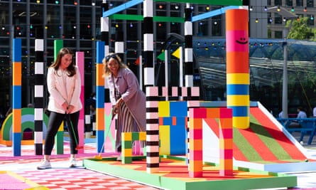 two women play on a brightly-coloured minigolf course with geometric shapes, stripes and patterns, with office buildings in the background