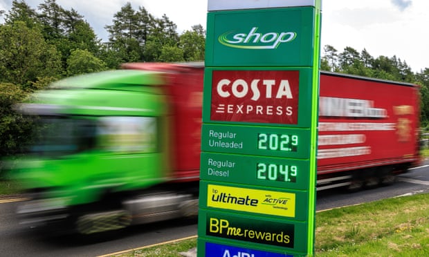 A green petrol station sign showing unleaded at £2.02 a litre and diesel at £2.04, with a large green and red lorry going past in a blur behind it