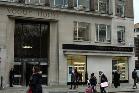 People walk past the frontage of Vogue House, a stone building with an imposing glass doorway and revolving doors. The scene appears wintry with the passersby wearing hats, coats and scarves, and many are holding large shopping bags.