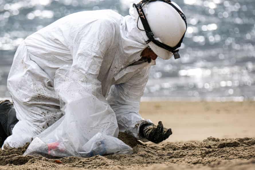A worker in a protective suit cleans up Huntington Beach, bending over the sand