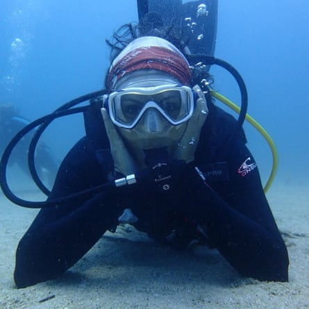 A person in diving gear looks to the camera with their arms resting on the seabed