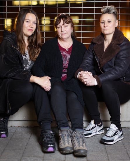 Three women sitting down together holding hands. They are all wearing dark jacket and trainers.