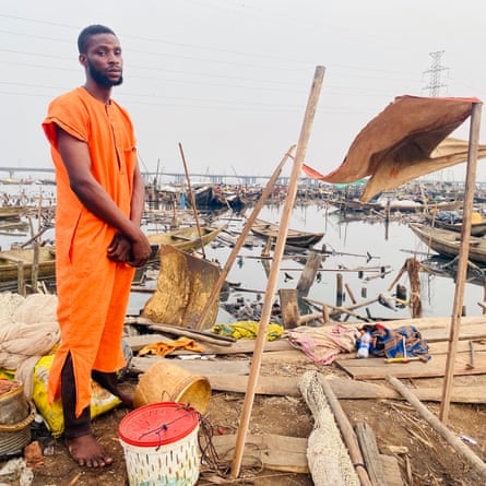 ‘They told us to leave. They didn’t tell us where to go’: the demolitions destroying homes and lives in Lagos A man in an orange jumpsuit stand next to a basic sunshade set up among the remains of his home