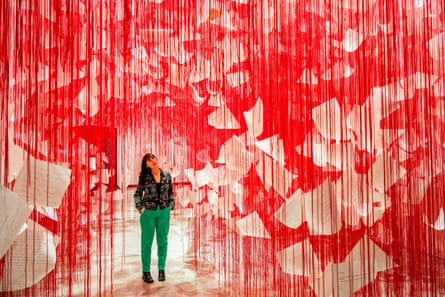 A woman stands below a sea of red threads hung with paper sheets.