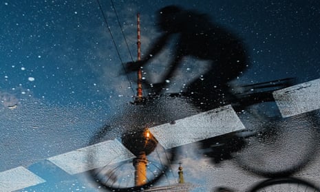 A woman riding a bicycle and the Berliner Fernsehturm reflected in a puddle on the street.