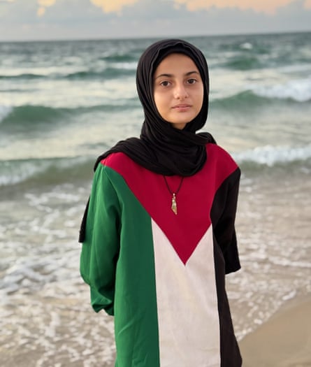 A girl wearing a black headscarf and a top decorated with the Palestinian flag stands on a shoreline looking at the viewer.