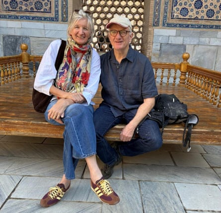 A man wearing a white cap, a blue shirt and jeans. A woman wearing a light blue shirt, jeans and brown and yellow trainers, and a floral scarf. They are sitting on a bench with a slatted wall and mosaics behind them