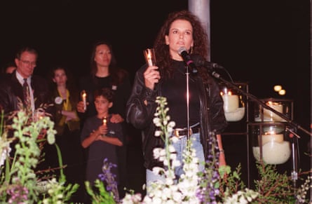 A woman with dark curly hair wearing a black jumper and blue jeans at a table holding a candle. Other people holding a candle are standing behind her
