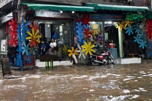 Two men sit in a shop whose front is decorated with brightly coloured propellers as floodwater flows by in the foreground