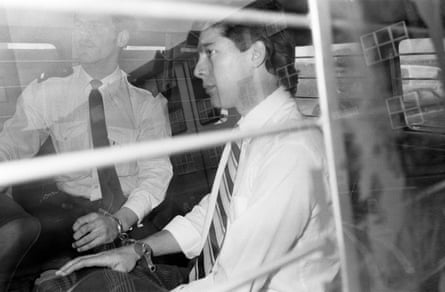 A man in a white shirt and stripy tie behind barriers in a police van with a policeman beside him.