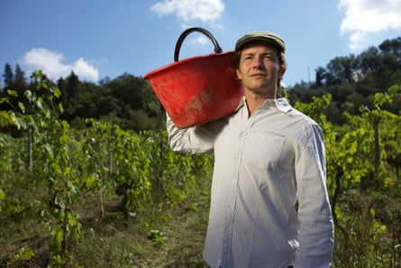 He stands in a vineyard carrying a large pannier (for grapes) on his shoulder, in the sun