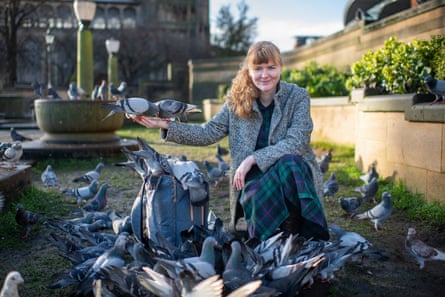 A woman kneels on the ground surrounded by pigeons as one eats out of her hand.