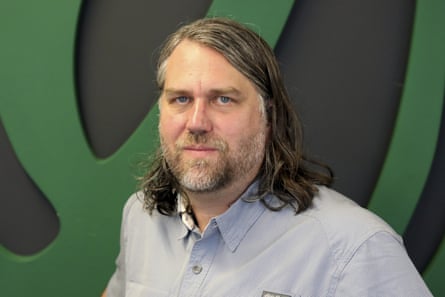 A portrait of Chad Nackers, the Onion’s editor-in-chief, a man with shoulder-length hair wearing a blue button-up shirt.