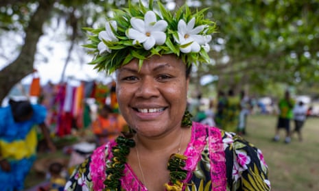 Julie King is smiling at the camera wearing a floral headdress and a brightly coloured shirt. Behind her are trees and people in a market