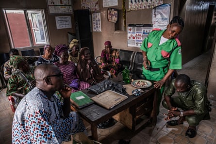 A group of men and women wearing African-print clothes sit and stand around a wooden desk in a room