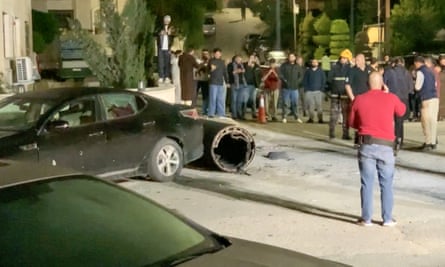About a dozen people stand around a large open cylinder lying next to the back of a car that has smashed windows