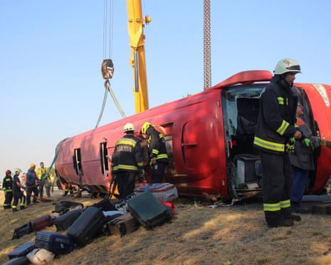 Firefighters at work next to the overturned bus, with suitcases lying on a slope next to it