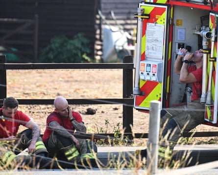 Two firefighters sitting on the roadside and another perched on the back of a fire engine