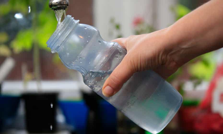 A hand refills a translucent reusable water bottle from a tap in an outdoor setting.