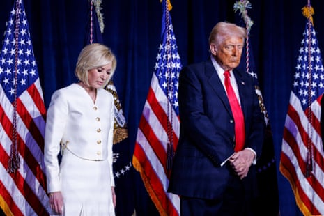a woman and a man stand with their heads bowed in front of American flags