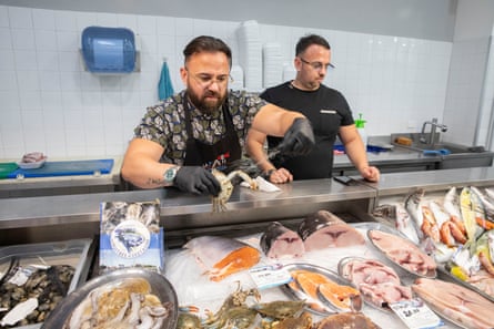 A fishmonger holds up a blue crab from a display of seafood, another man standing by his side.
