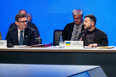 Man in glasses and suit on left, sitting at a table with man in patterned jumper on right, with other men in background.