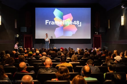 a group of people sit down and look ahead at a person standing on stage in front of theater screen