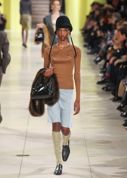 A model on a catwalk wearing a black cloche hat, a long-sleeved brown top, a silky skirt, knee socks and loafers, and carrying a handbag and a fur stole