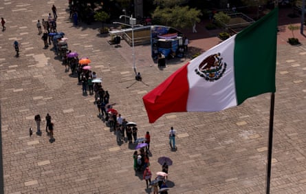An aerial view shows a line snaking across a plaza with the Mexican flag in the foreground.