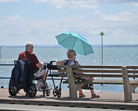 A man in a mobility scooter and a woman under a turquoise umbrella sit on a bench facing the sea