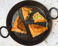 Four triangular golden-brown samosas sit in a black pan, one split open to reveal a vegetable filling.