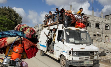 men and boys sit in a metal rack on the roof of a battered white van alongside bags of possessions and gas cylinders, while another man leans out of the van's side door. They are driving down a street of ruined buildings and the back of a motorcycle laden with brightly coloured bedding and a gas cylinder can be seen in the foreground.