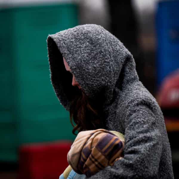 A woman in a hooded coat that obscures her face carries a bundle