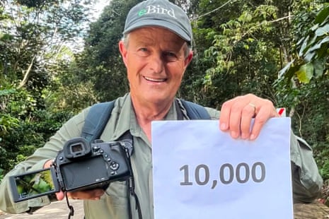 An older white man wearing an olive button-up shirt and a gray hat that says eBird, smiles amid trees as he holds in his right hand a digital camera with the back opened to show the image of a bird, and in his right hand a print-out of the number 10,000.