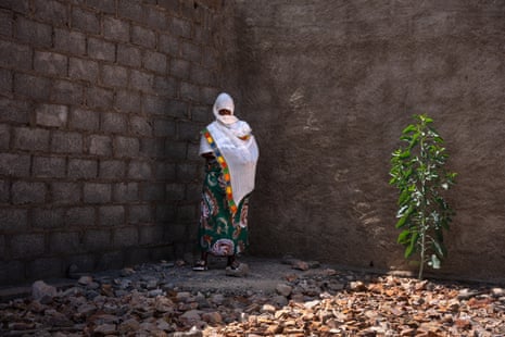 A woman, her head and fac wrappend ina white scarf, stands in a corner formed by two high cinder block walls. The ground around her is covered with leaves and a plant grows beside her.