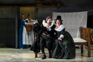 A man and woman in period costume sit on stage together on a bed with two women dressed as maids behind