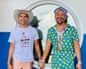 Ben Graetz and Dion Padan at a boat party in Darwin. Ben is wearing a broad brim sun hat and white Darwin Pride festival T-shirt. Dion is in a sailor hat, a blue button shirt with yellow rubber ducks that is unbuttoned to his navel. They look happy and are holding hands.