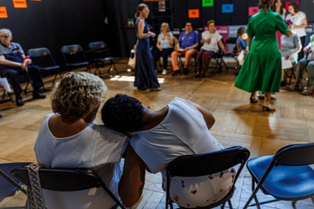 Participants in Escuela de Espectadores Sénior rehearse plays being shown at the Matadero arts centre. In the foreground, one seated woman rests her head on another’s shoulder; in the distance, two actors rehearse their performance, before more participants in the programme
