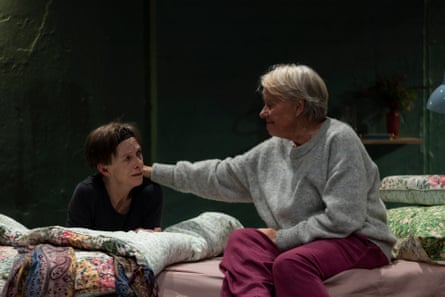 Two middle-aged women sat on a mattress with one placing her arm on the other’s shoulder.